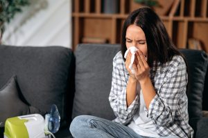 Picture of a woman sneezing into a tissue while sitting on a couch.